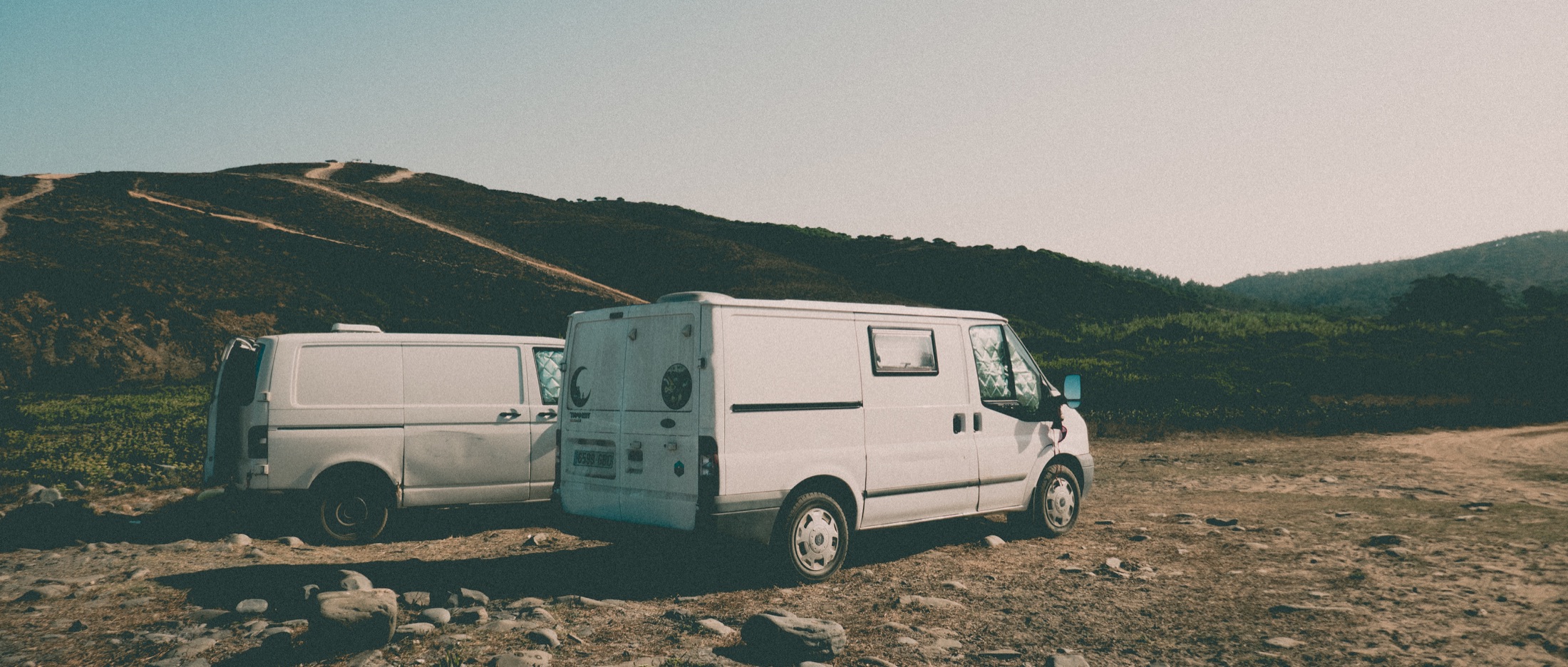 Two white camper vans parked beneath coastal hills
