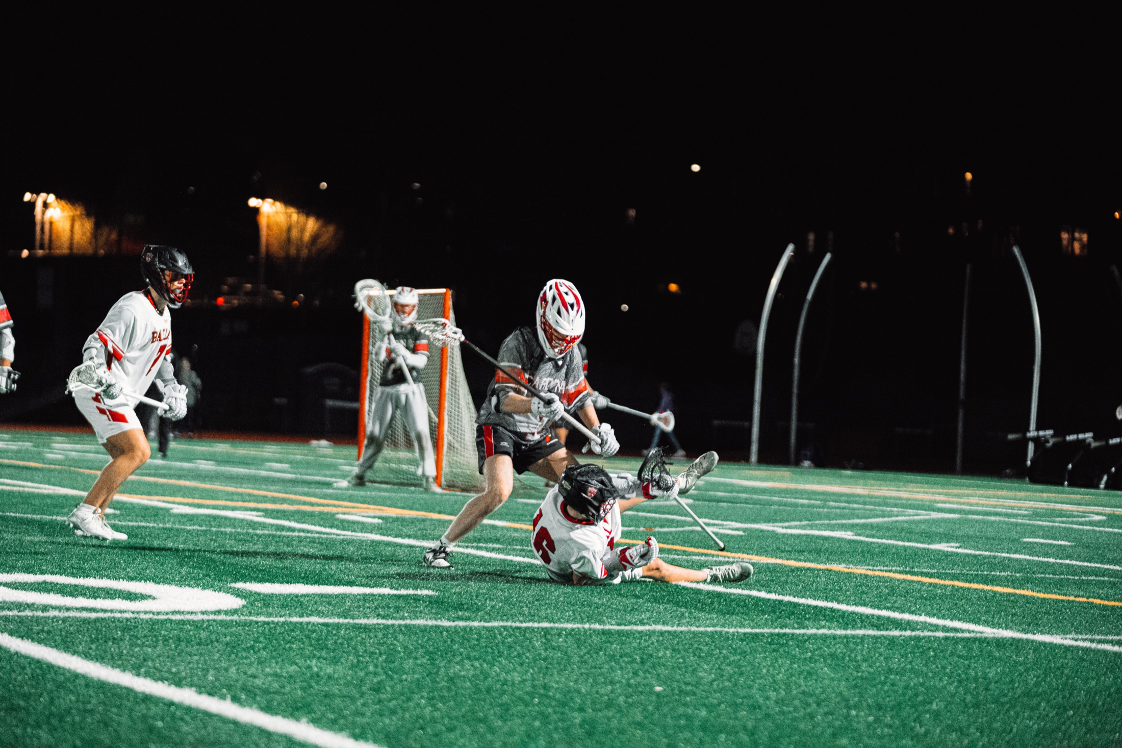 Night lacrosse action as a ball carrier falls under a defensive check