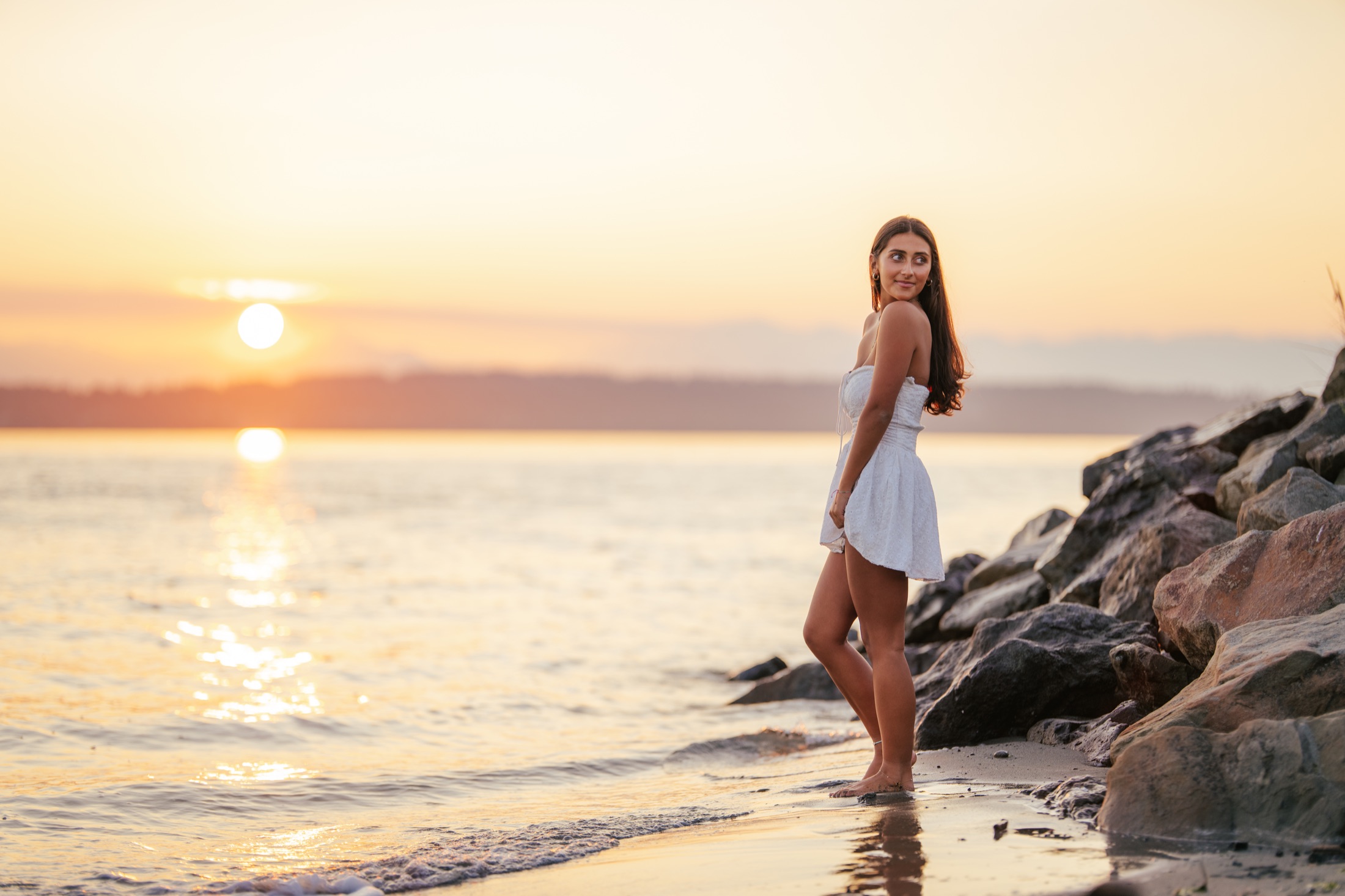 Young woman in a white dress standing beside the water at sunset