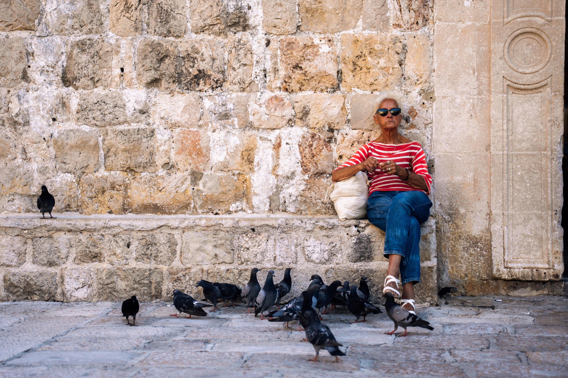 Woman in a striped shirt seated against a stone wall among pigeons